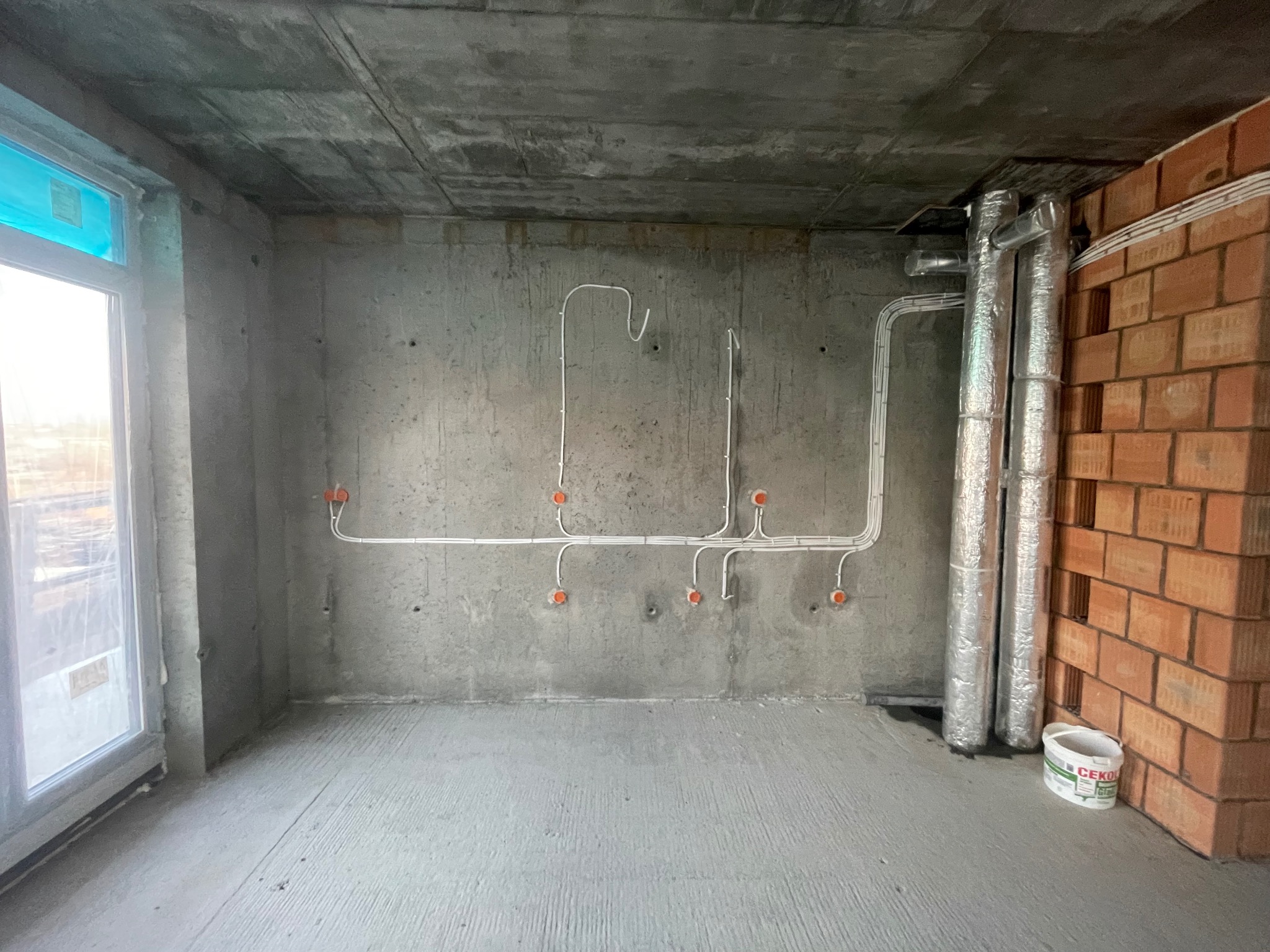 Surowe wnętrze w trakcie wykańczania: widoczne przewody elektryczne poprowadzone po betonowej ścianie, fragment instalacji wentylacyjnej w srebrnej izolacji oraz mur z czerwonej cegły. Na podłodze...