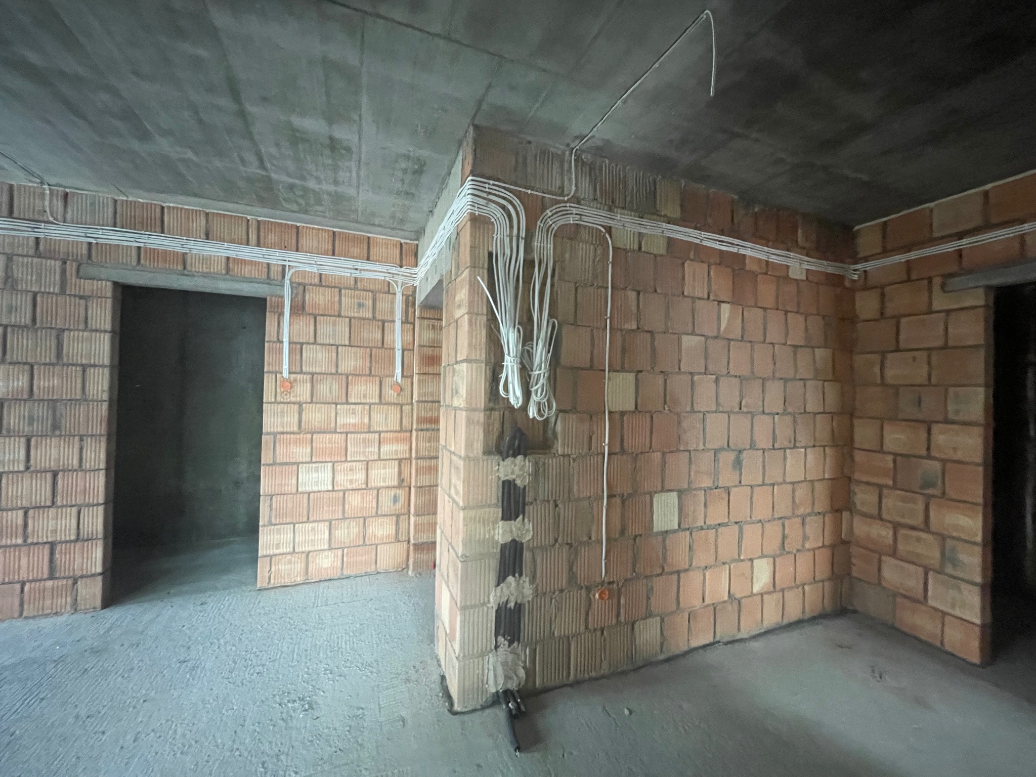 Surowy stan wnętrza z widocznymi przewodami elektrycznymi ułożonymi na ścianach z cegły i pod sufitem z betonu.