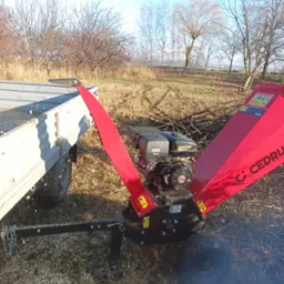 Czerwony rębak do gałęzi marki Cedrus podłączony do przyczepki samochodowej na tle łąki z drzewami bez liści, w otoczeniu wiórów drzewnych.