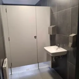 Wnętrze publicznej toalety z kabinami z szarego laminatu, umywalką na tle ciemnych płytek i grzejnikiem na ścianie.