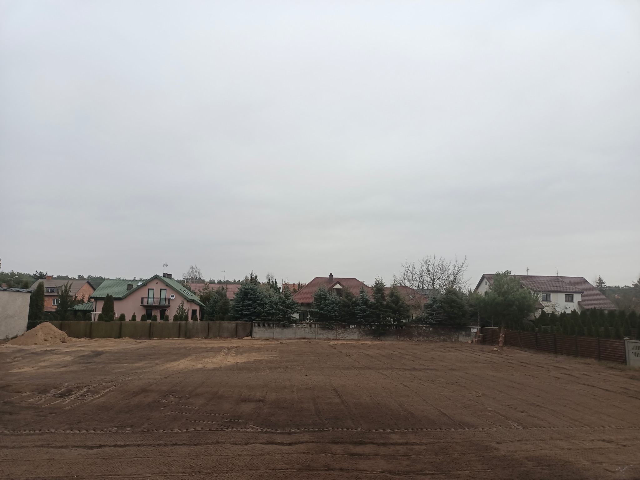 Przygotowany teren pod budowę domu, świeżo wyrównana ziemia z widocznymi śladami opon, w tle zabudowa jednorodzinna i pochmurne niebo.