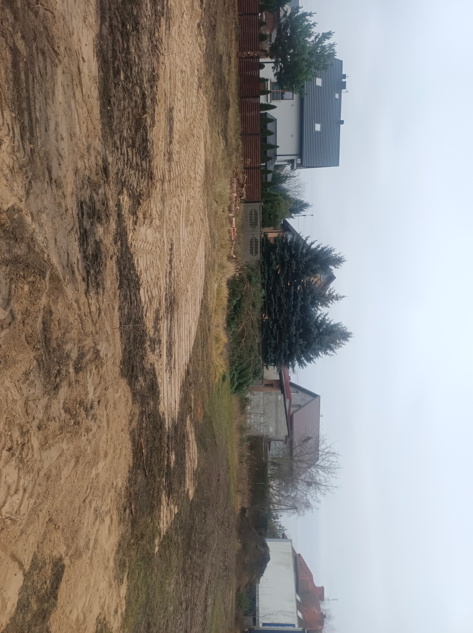 Przygotowany teren pod budowę z widocznymi śladami opon, sąsiednie domy w tle.