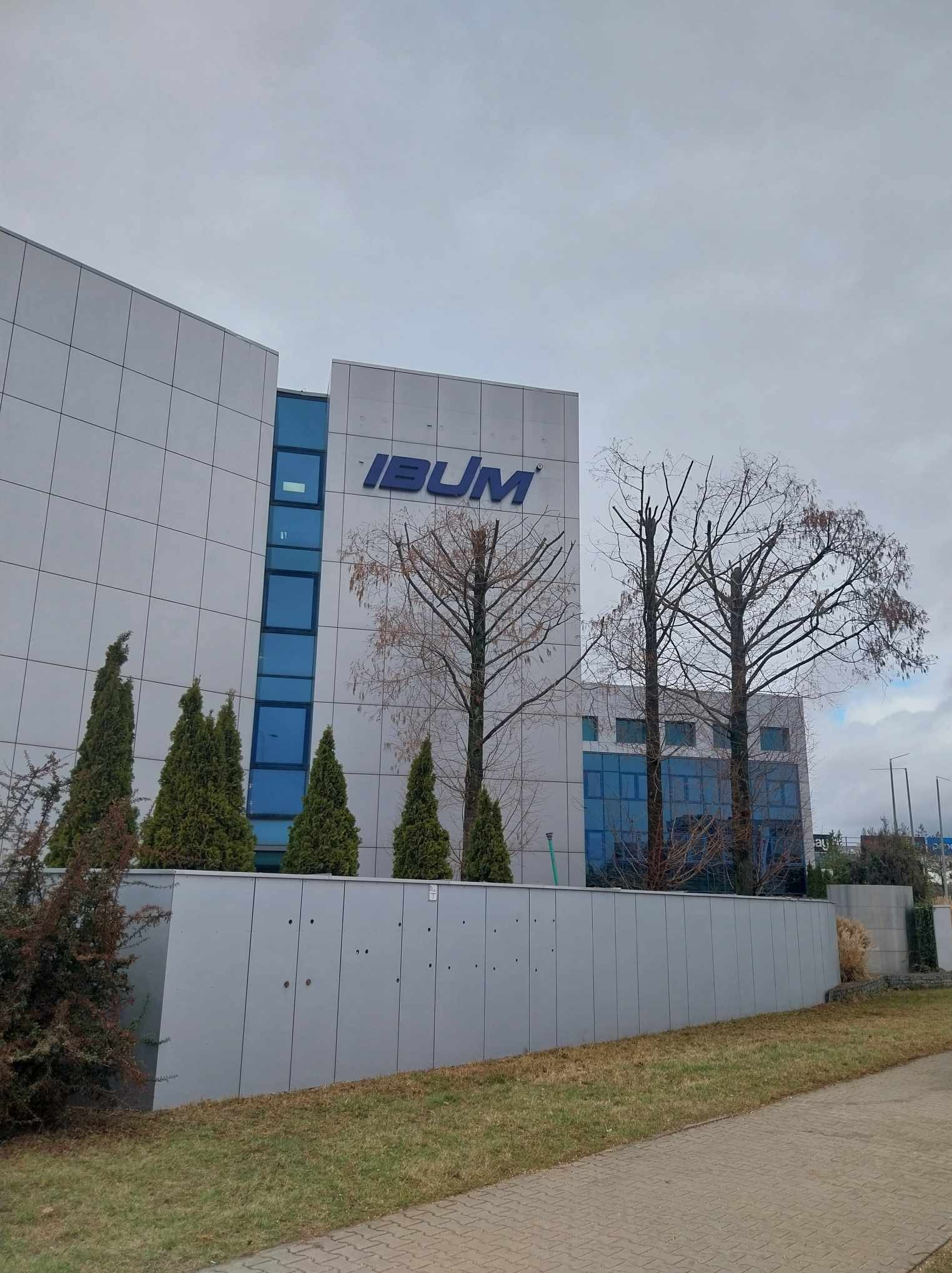 Nowoczesny budynek biurowy z logo IBUM, otoczony zielenią i drzewami po przycince, widok z poziomu chodnika.