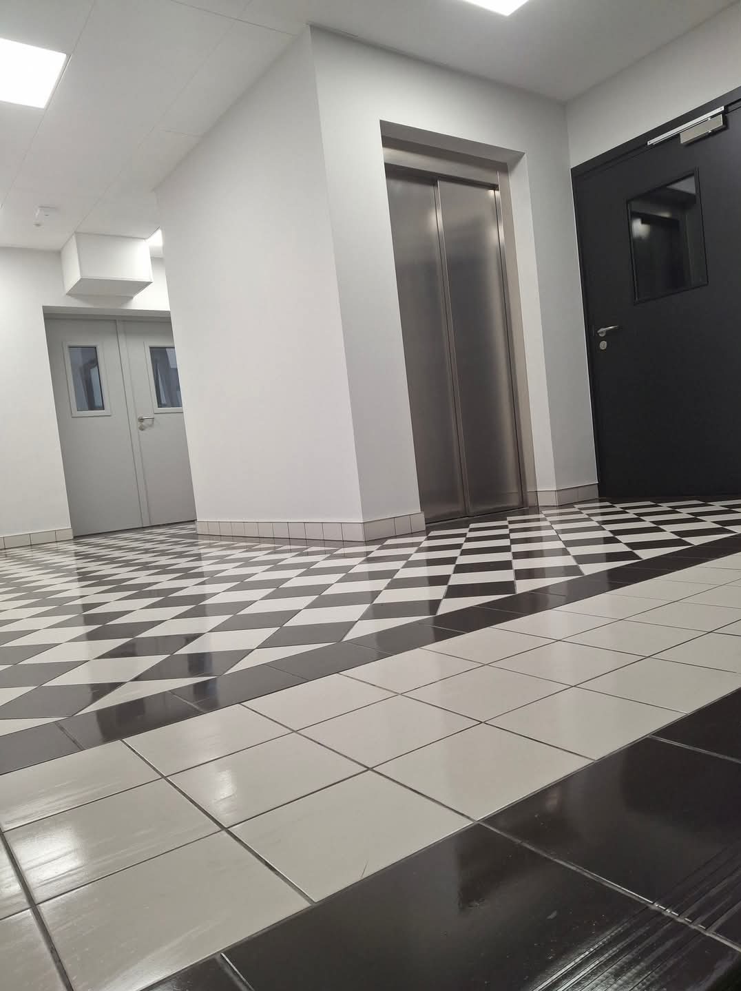 Błyszcząca, czarno-biała szachownica płytek podłogowych w korytarzu, z widocznymi drzwiami windy i drzwiami wejściowymi, oświetlonymi przez górne światło.