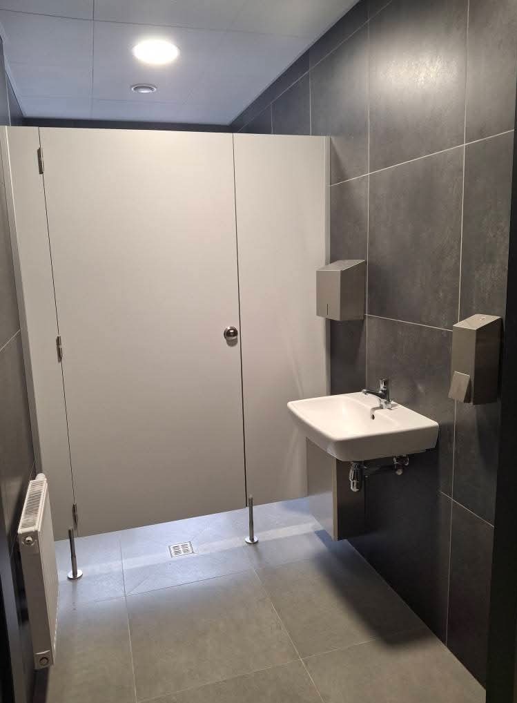 Wnętrze publicznej toalety z kabinami z szarego laminatu, umywalką na tle ciemnych płytek i grzejnikiem na ścianie.