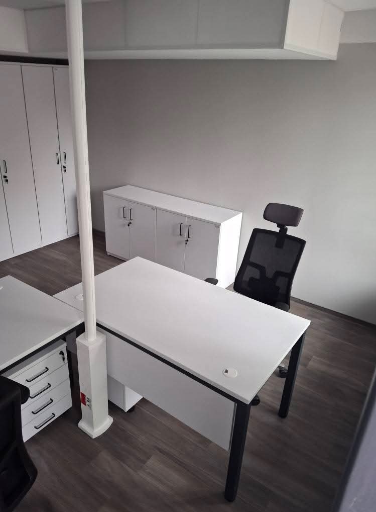 Nowoczesne biuro z białymi meblami: biurka z czarnymi nogami, ergonomiczne krzesło, szafy zamykane na klucz, drewnopodobna podłoga, oświetlenie sufitowe.