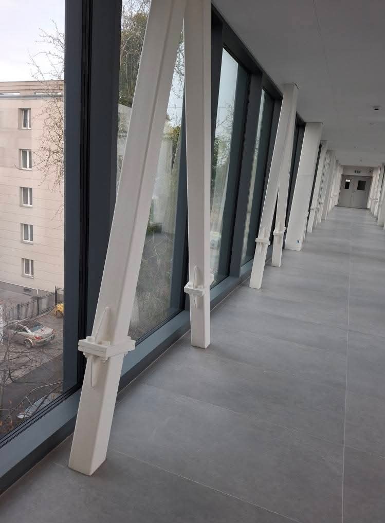 Wnętrze korytarza z przeszkloną ścianą wspartą na skośnych, białych podporach, widok na budynek i parking na zewnątrz.