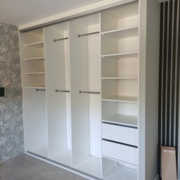 Lamit-Meble Jacek Andrzejczyk - Jasna szafa wnękowa z przesuwnymi drzwiami, półkami i szufladami. Widoczne drążki na ubrania. Na ścianie dekoracyjne panele pionowe i tapeta we wzory.