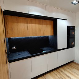 Lamit-Meble Jacek Andrzejczyk - Nowoczesna kuchnia na wymiar: białe fronty szafek dolnych i górnych, drewniane szafki wiszące, czarny blat i ściana nad blatem, zabudowane piekarniki.