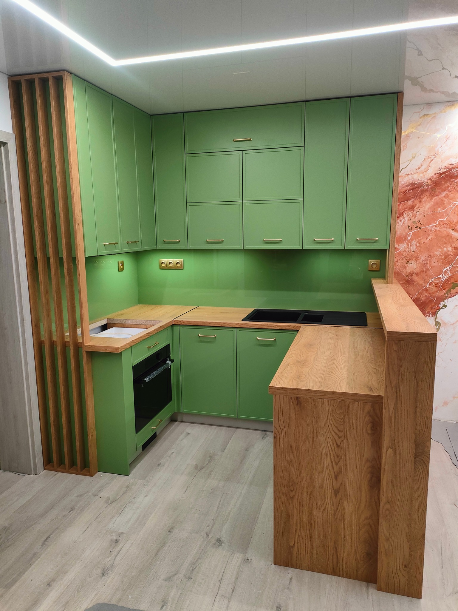 Nowoczesna, jasnozielona zabudowa kuchenna z drewnianym blatem i barkiem, oświetleniem LED podszafkowym i dekoracyjną ścianą w odcieniach czerwieni i złota.