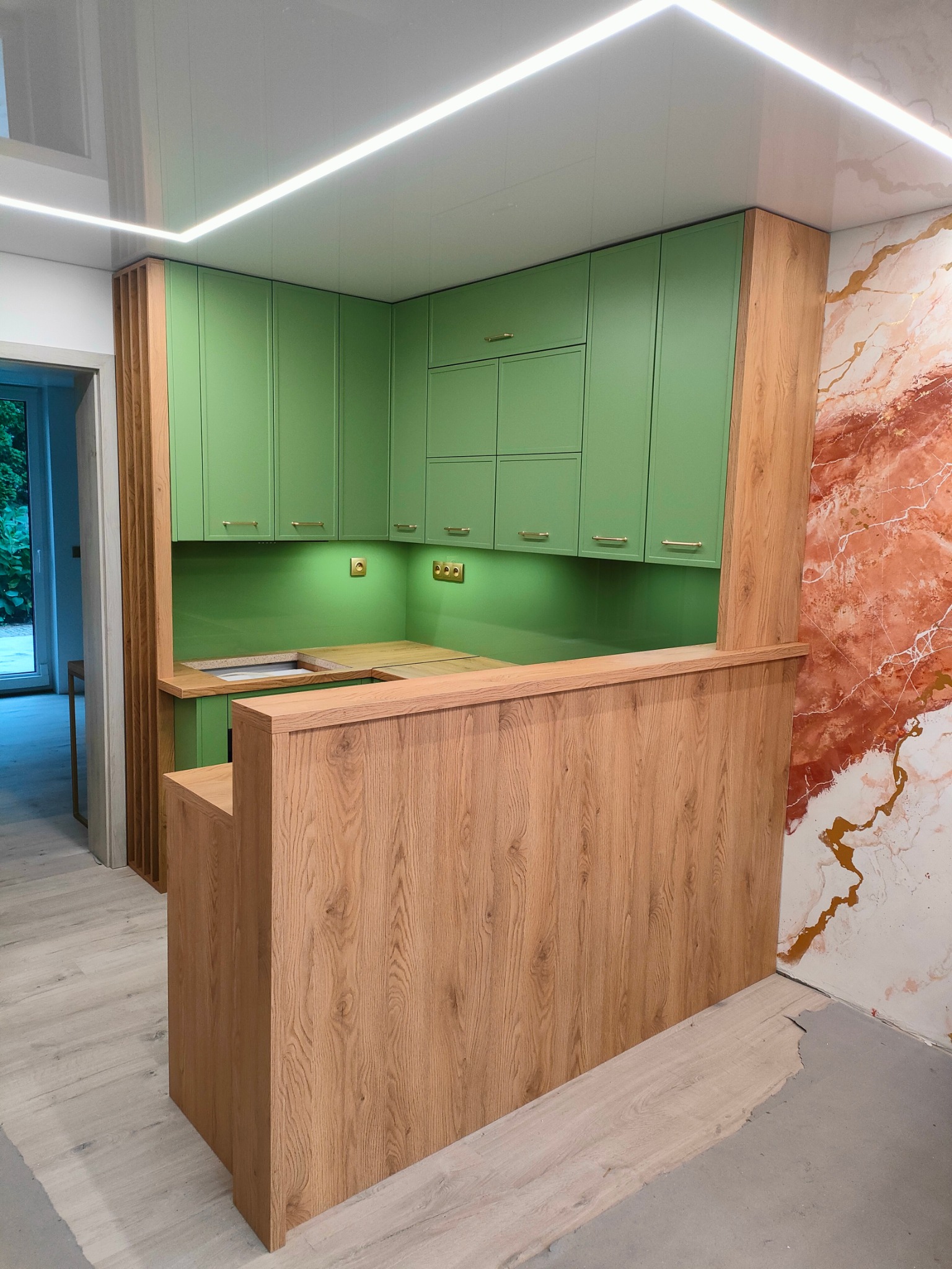 Nowoczesna kuchnia z zielonymi szafkami i drewnianym blatem, widoczne gniazdka elektryczne na ścianie, fragment ściany z efektem marmuru, sufit z oświetleniem LED.