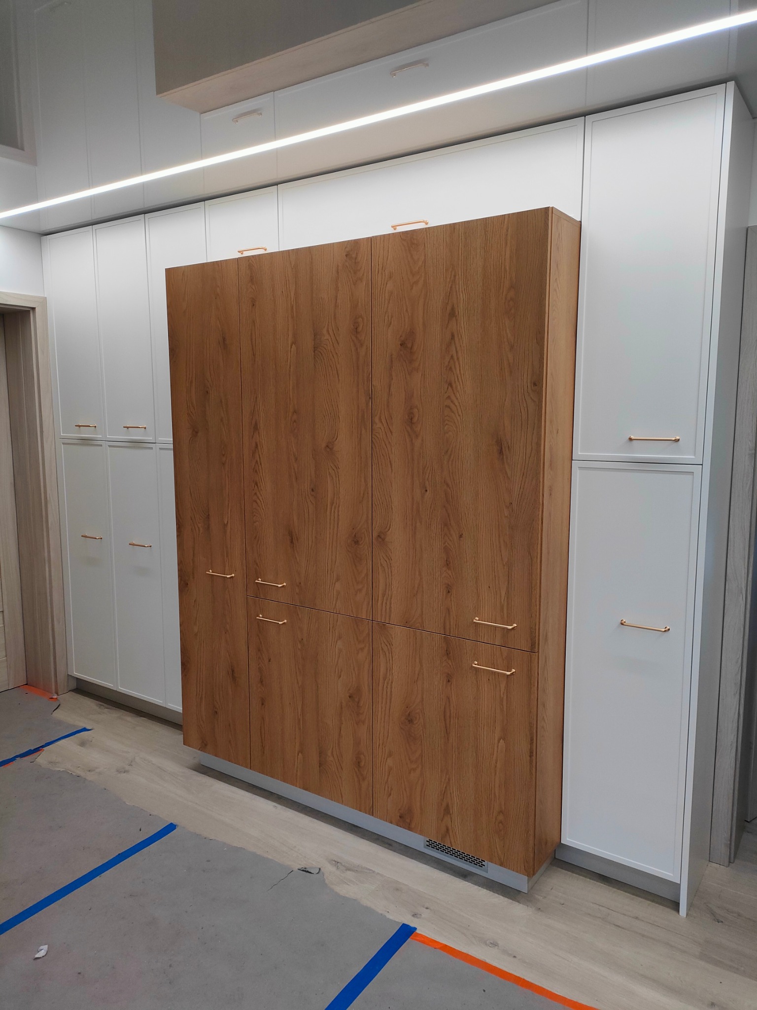 Zabudowa wnękowa: wysokie szafy z białymi frontami i drewnopodobnym panelem centralnym, podłużne złote uchwyty, oświetlenie LED podszafkowe, widoczne ślady prac wykończeniowych na podłodze.