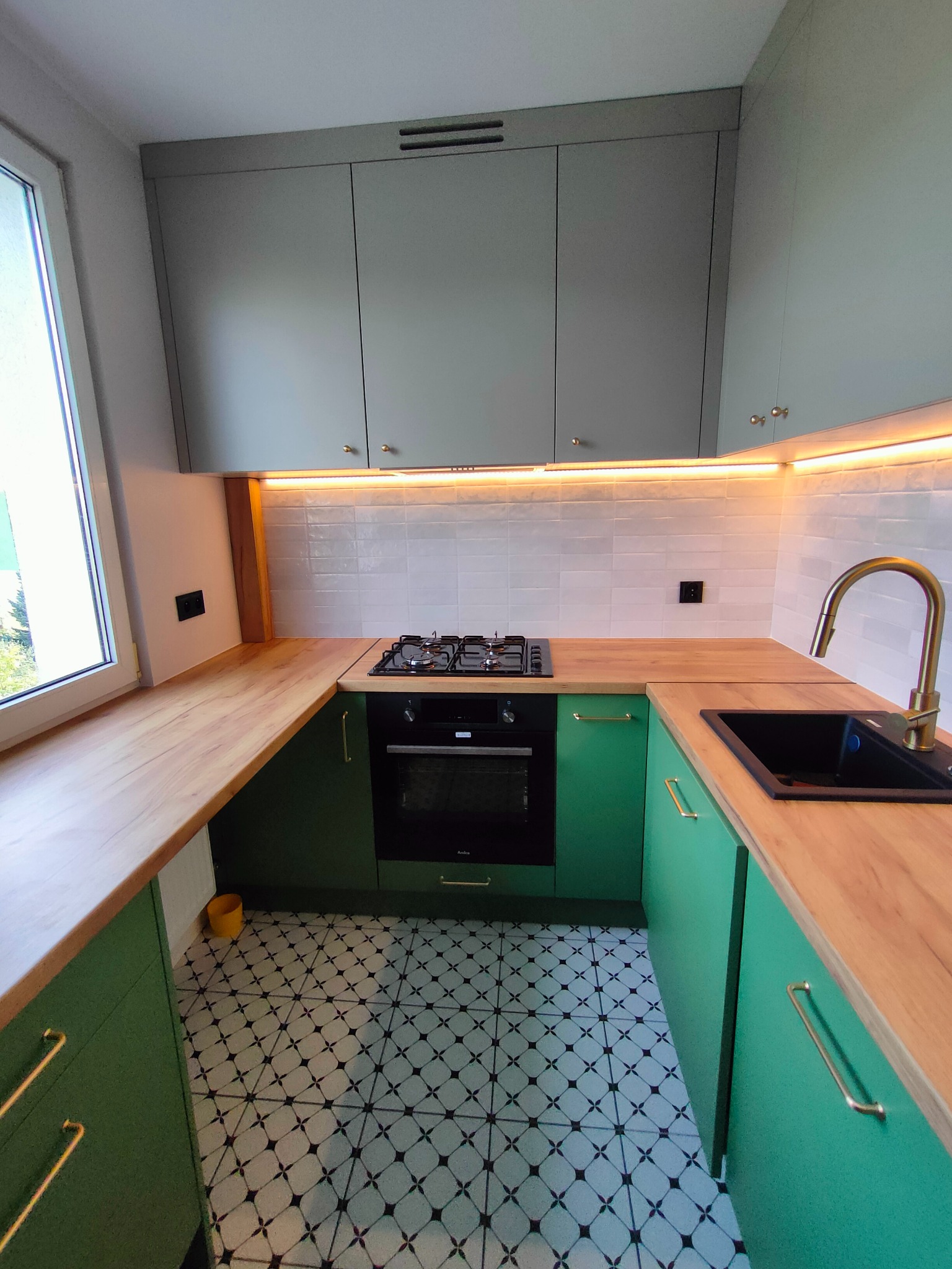 Nowoczesna kuchnia w odcieniach zieleni i szarości z drewnianym blatem, oświetleniem LED pod szafkami wiszącymi i czarnym zlewem z baterią w kolorze złotym.