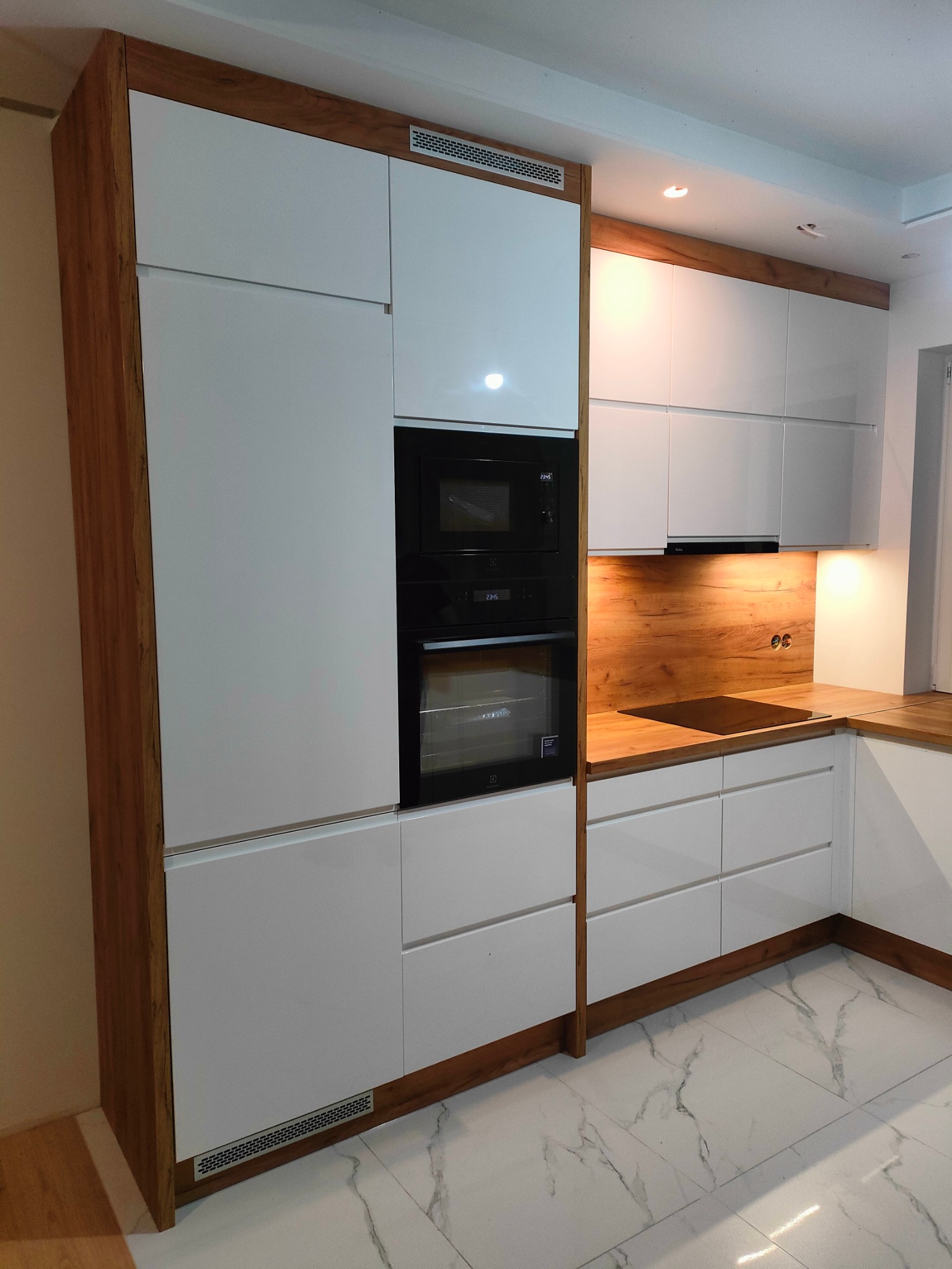 Nowoczesna kuchnia na wymiar w Gomunicach: białe fronty szafek z drewnianymi akcentami, zabudowany piekarnik i mikrofalówka, marmurowe płytki podłogowe.