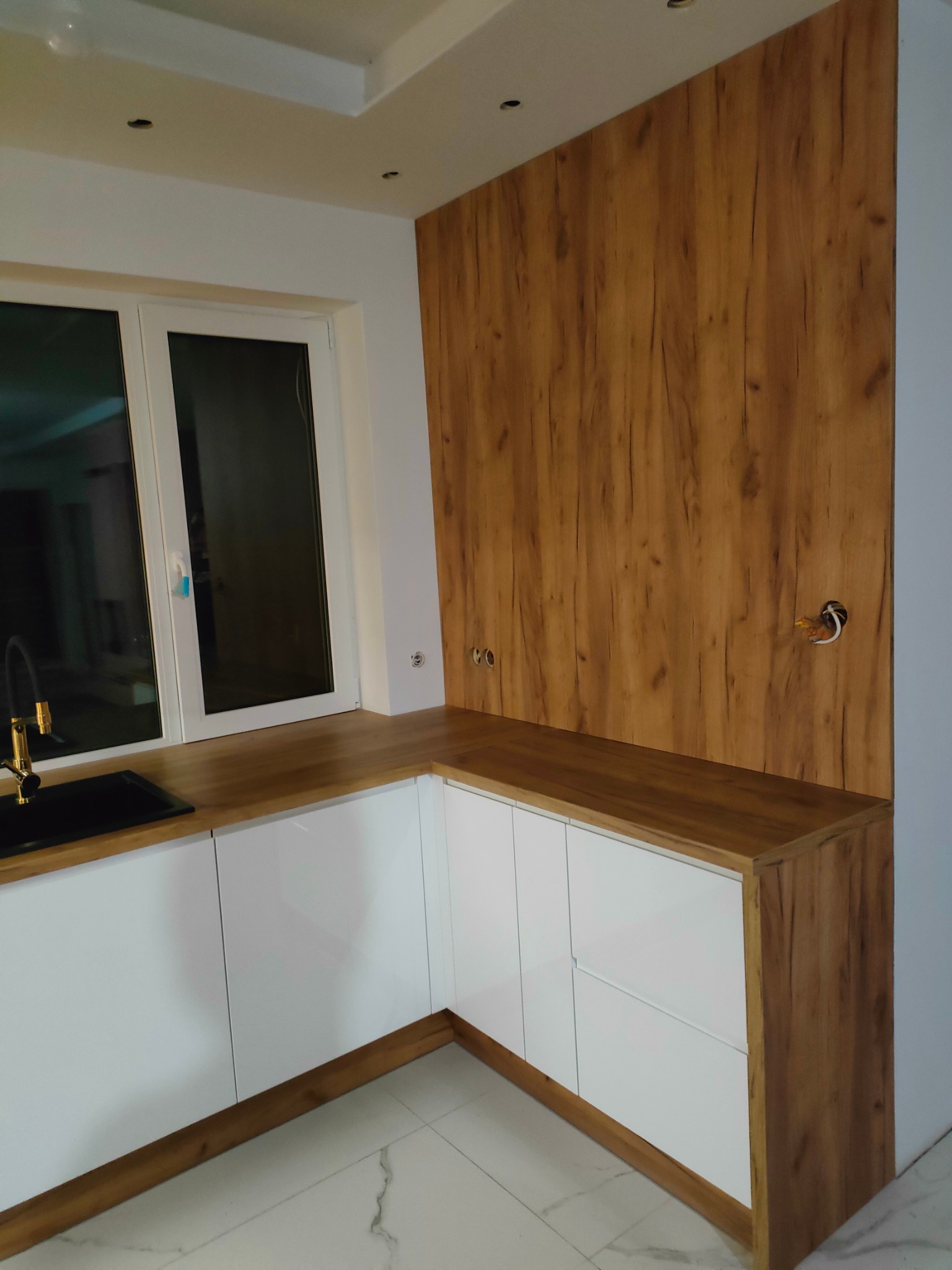 Nowoczesna kuchnia z białymi frontami szafek i drewnianym blatem oraz panelem ściennym o strukturze drewna, widoczne gniazdka elektryczne w ścianie.