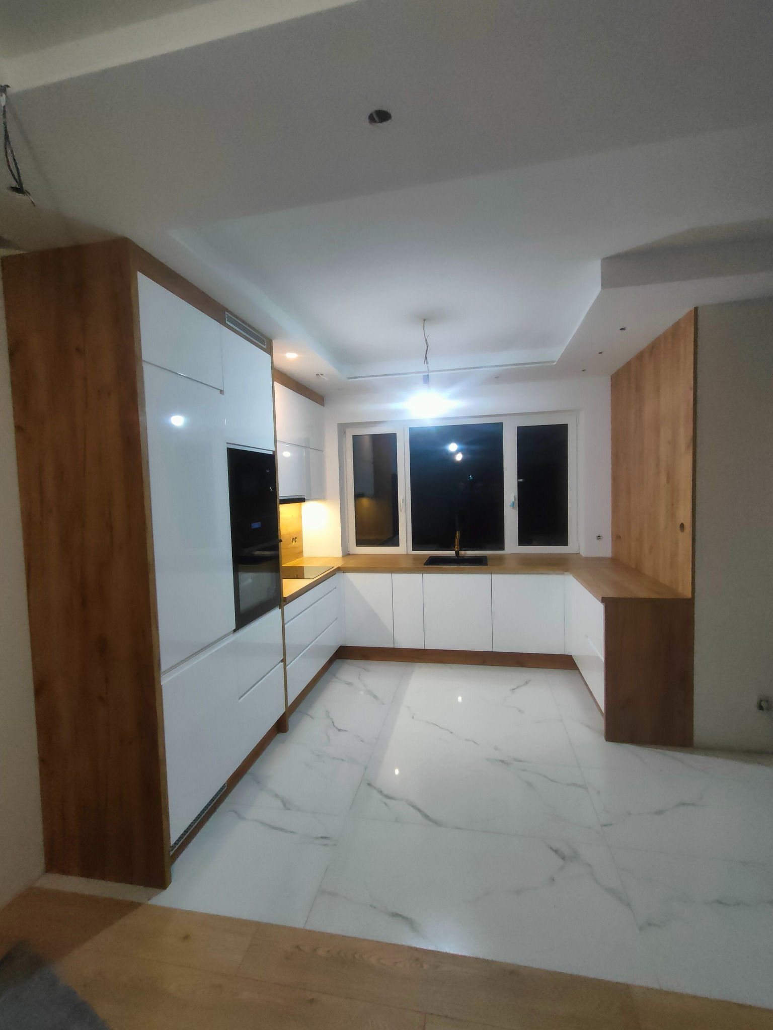 Nowoczesna kuchnia z białymi frontami i drewnianymi akcentami, marmurowa podłoga, widoczne niedokończone prace elektryczne na suficie.