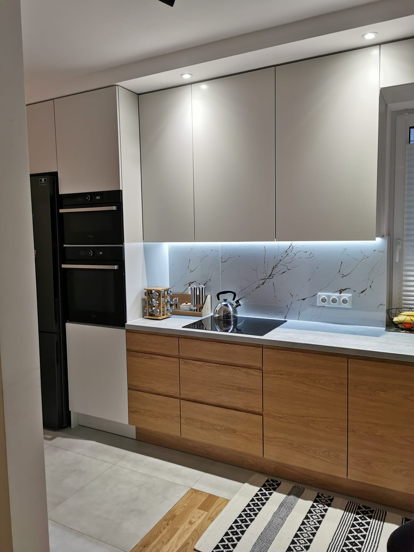 Nowoczesna kuchnia z drewnianymi frontami szafek dolnych, białymi szafkami górnymi, marmurowym backsplashiem z podświetleniem LED, czarnym piekarnikiem i lodówką oraz czajnikiem na płycie indukcyjnej.