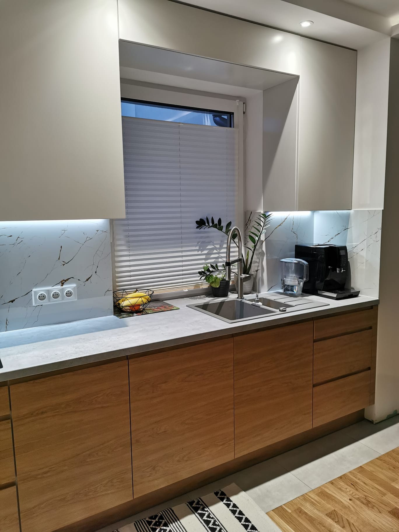 Nowoczesna kuchnia z drewnianymi frontami szafek dolnych i białymi górnymi, marmurowy backsplash z podświetleniem LED, stalowy zlew z baterią, rolety w oknie, ekspres do kawy i rośliny doniczkowe...