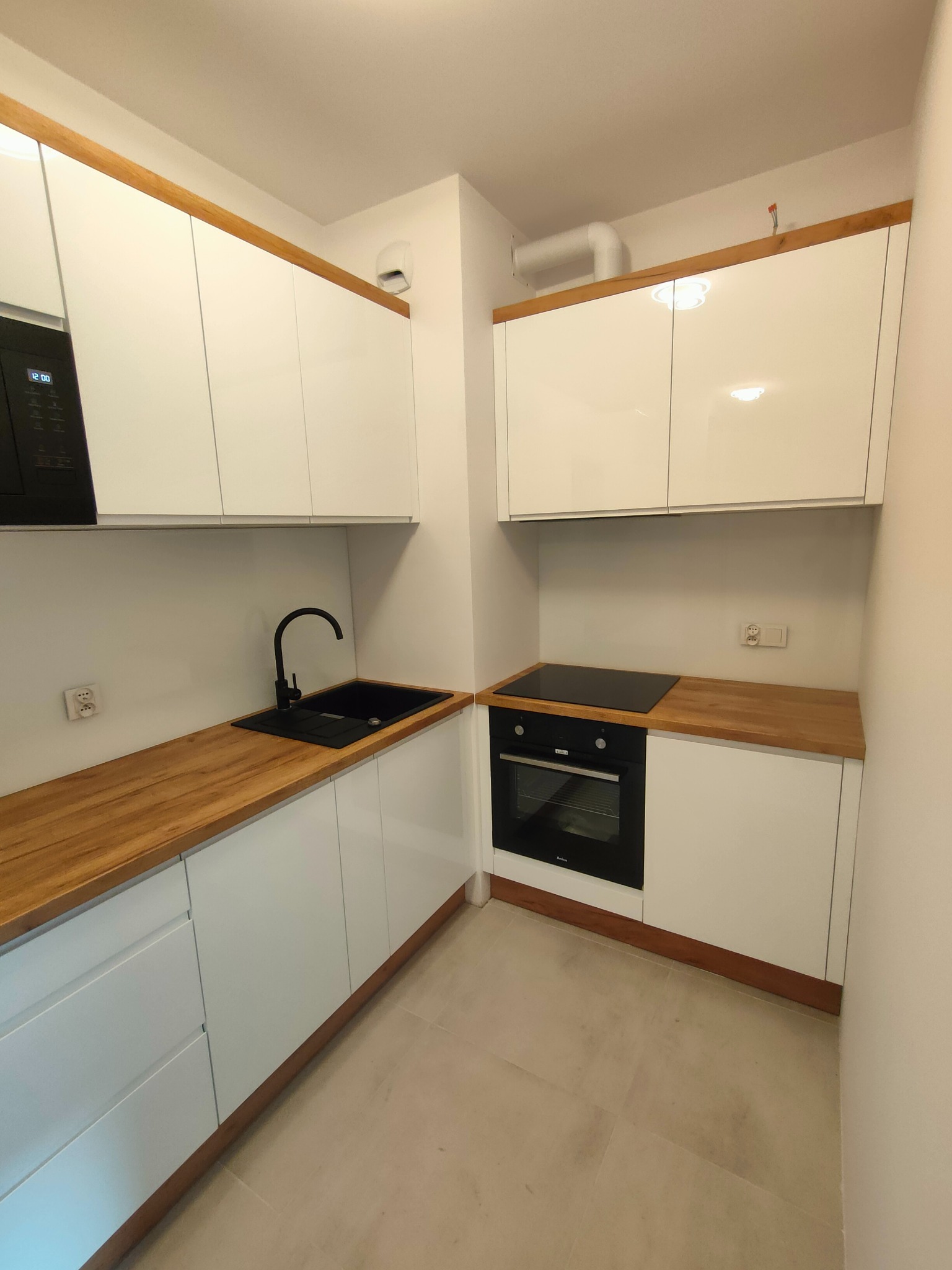 Nowoczesna, kompaktowa kuchnia z białymi frontami, drewnianym blatem i czarną armaturą, oświetlona lampami halogenowymi w górnych szafkach.