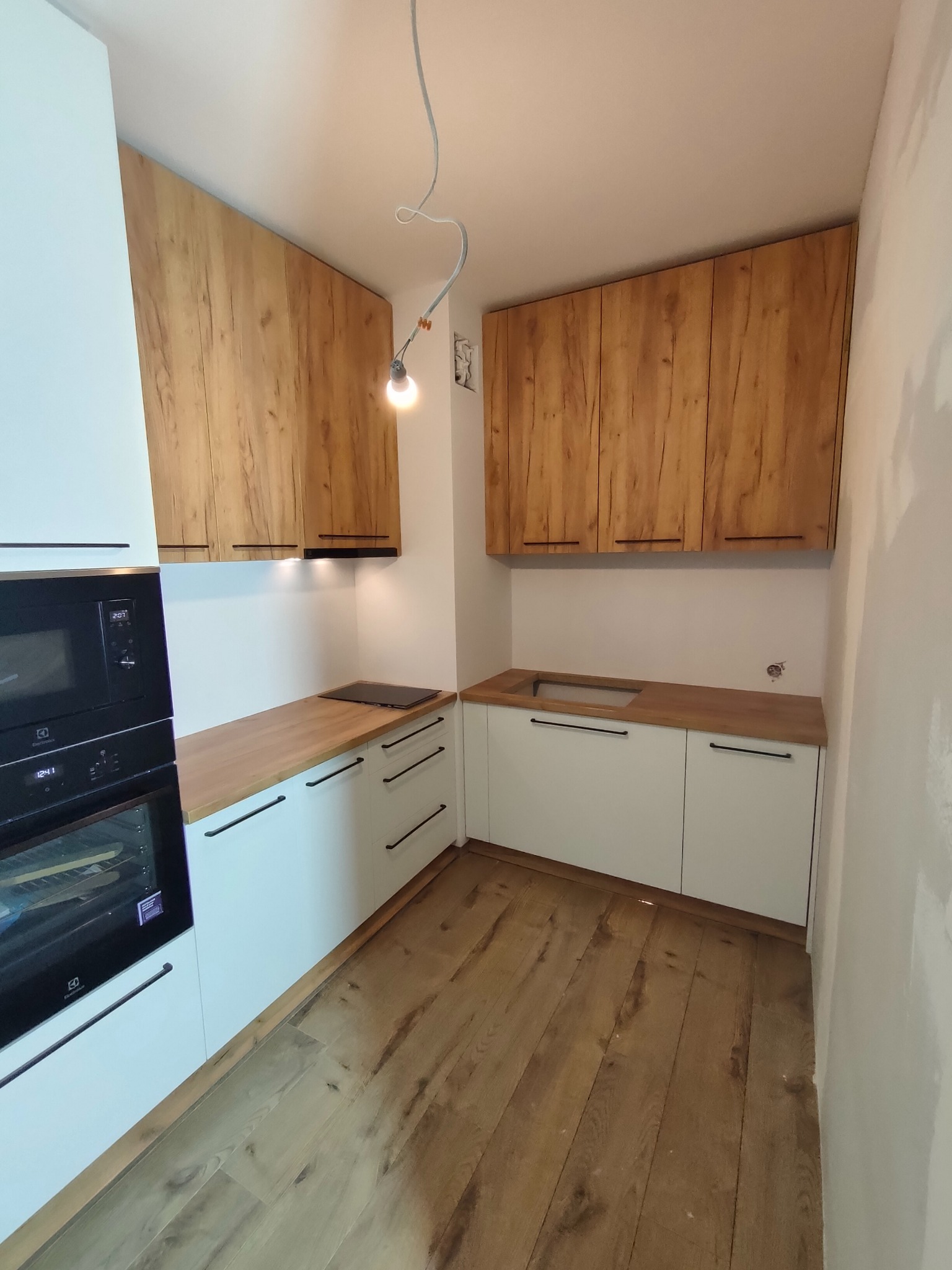 Nowoczesna kuchnia z białymi szafkami dolnymi i górnymi szafkami z drewnopodobnym fornirem, czarne uchwyty, drewniana podłoga, widoczne niedokończone ściany.