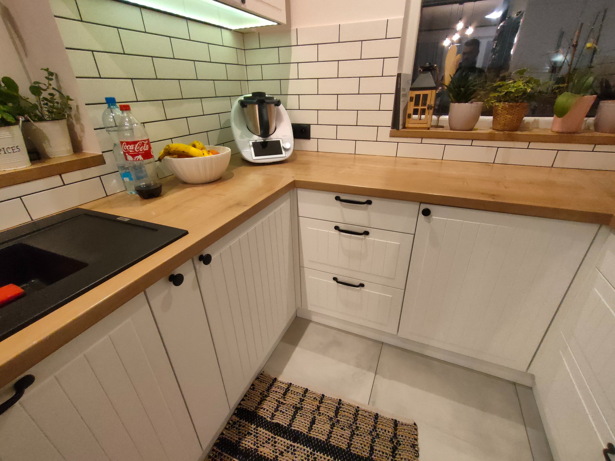 Jasna kuchnia z drewnianym blatem, białymi szafkami z frezowanymi frontami i czarnymi uchwytami, widok na okno z roślinami doniczkowymi i robot kuchenny na blacie