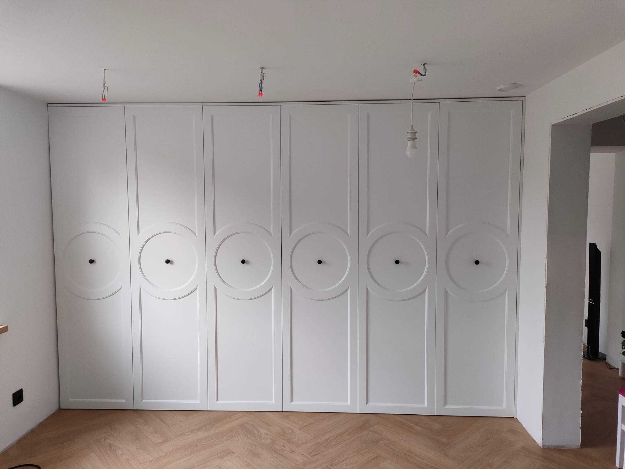 Biała, zabudowana szafa wnękowa z sześcioma drzwiami, każdy z okrągłym, dekoracyjnym elementem i czarną gałką, na podłodze parkiet ułożony w jodełkę, widoczne niedokończone prace elektryczne...