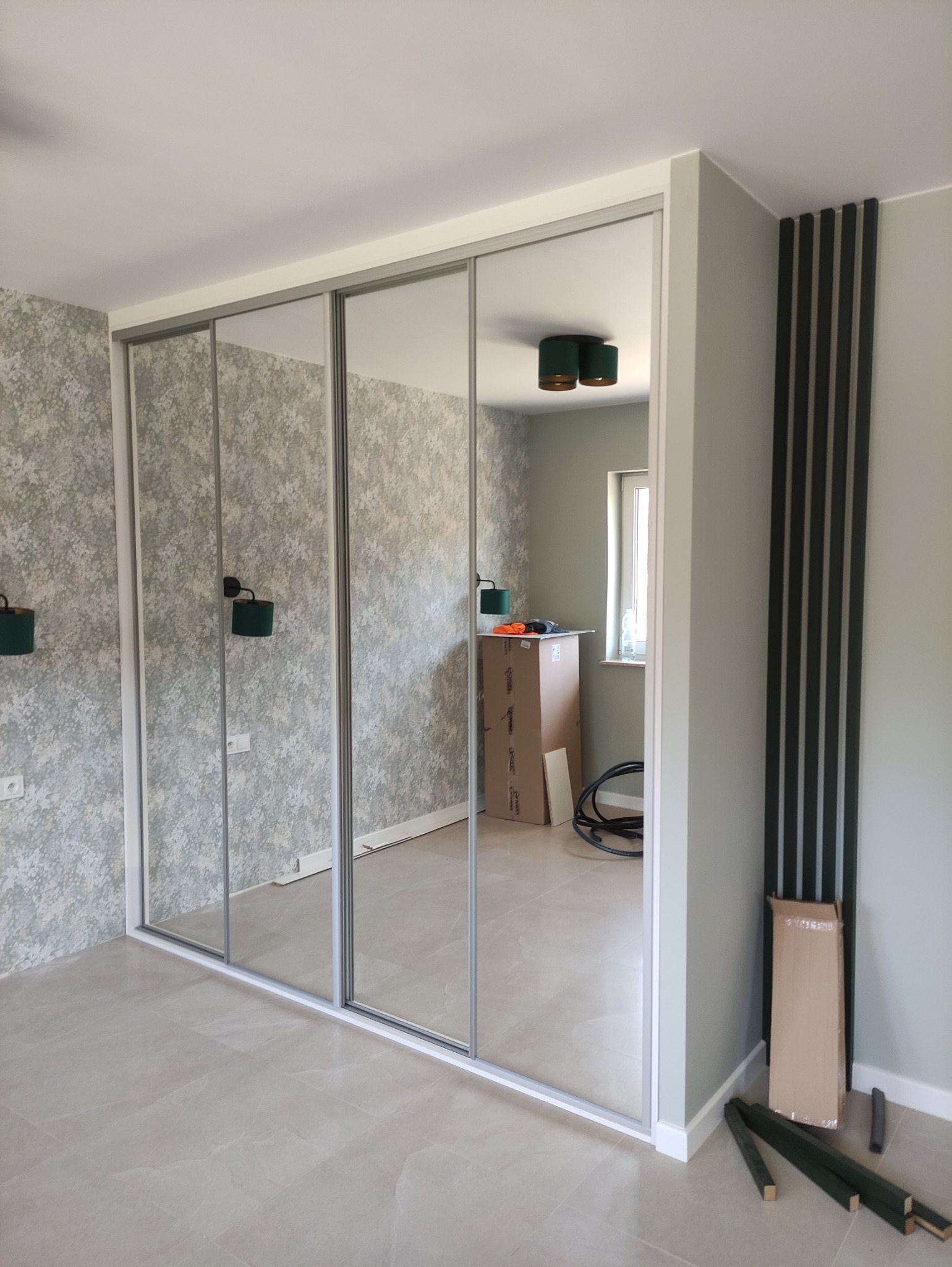 Jasne wnętrze z szafą wnękową z lustrzanymi drzwiami przesuwnymi. Widoczne dekoracyjne panele ścienne i kartonowe pudło w pomieszczeniu.