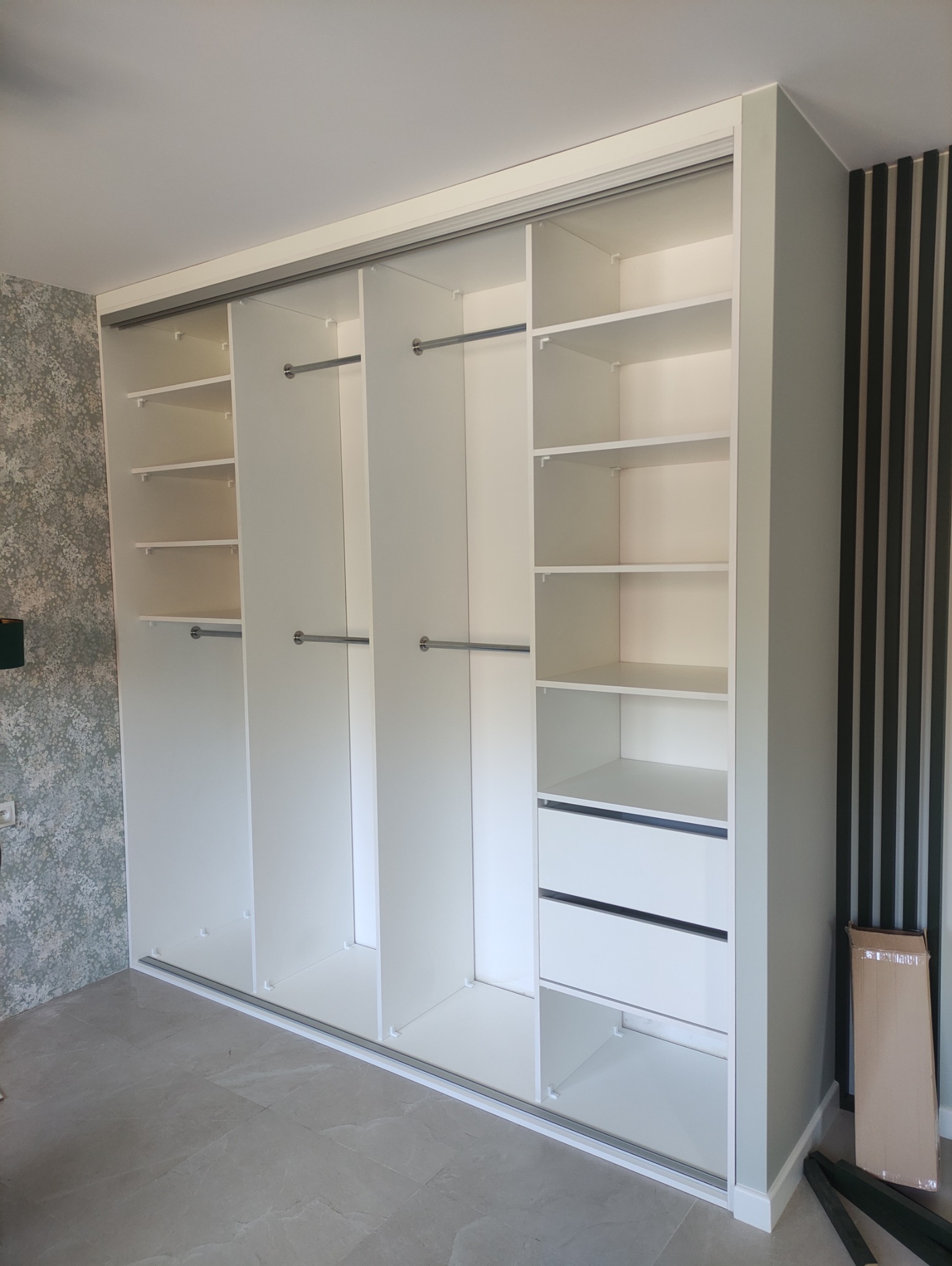 Jasna szafa wnękowa z przesuwnymi drzwiami, półkami i szufladami. Widoczne drążki na ubrania. Na ścianie dekoracyjne panele pionowe i tapeta we wzory.