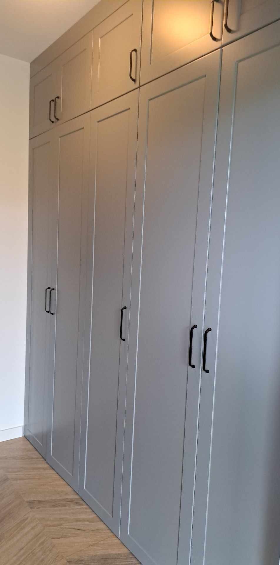Szara, zabudowana szafa z czarnymi uchwytami, dopasowana do podłogi z paneli w jodełkę. Górne szafki sięgają sufitu. Minimalistyczny design.