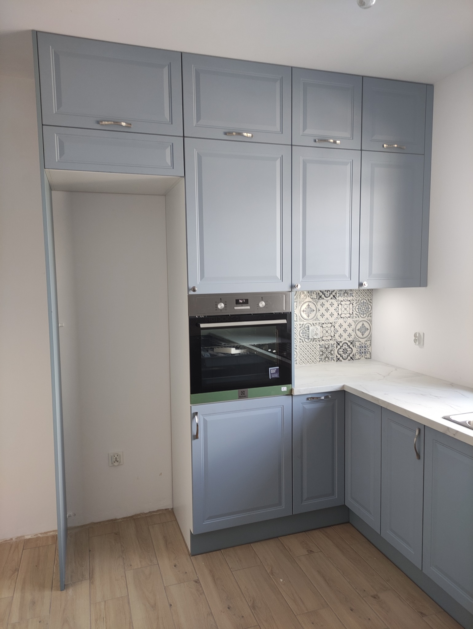 Niebieska kuchnia z piekarnikiem i wzorzystymi płytkami. Górne szafki wiszące z srebrnymi uchwytami. Podłoga wyłożona drewnopodobnymi panelami.