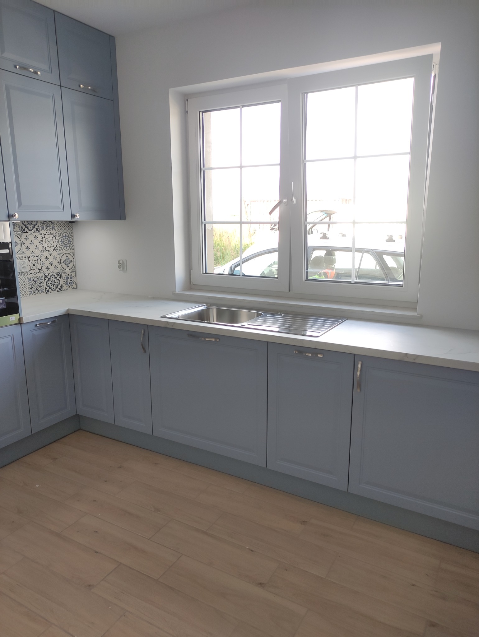 Jasnoszara kuchnia z frezowanymi frontami, białym blatem i wzorzystymi płytkami nad zlewem, oświetlona przez okno z widokiem na zewnątrz.