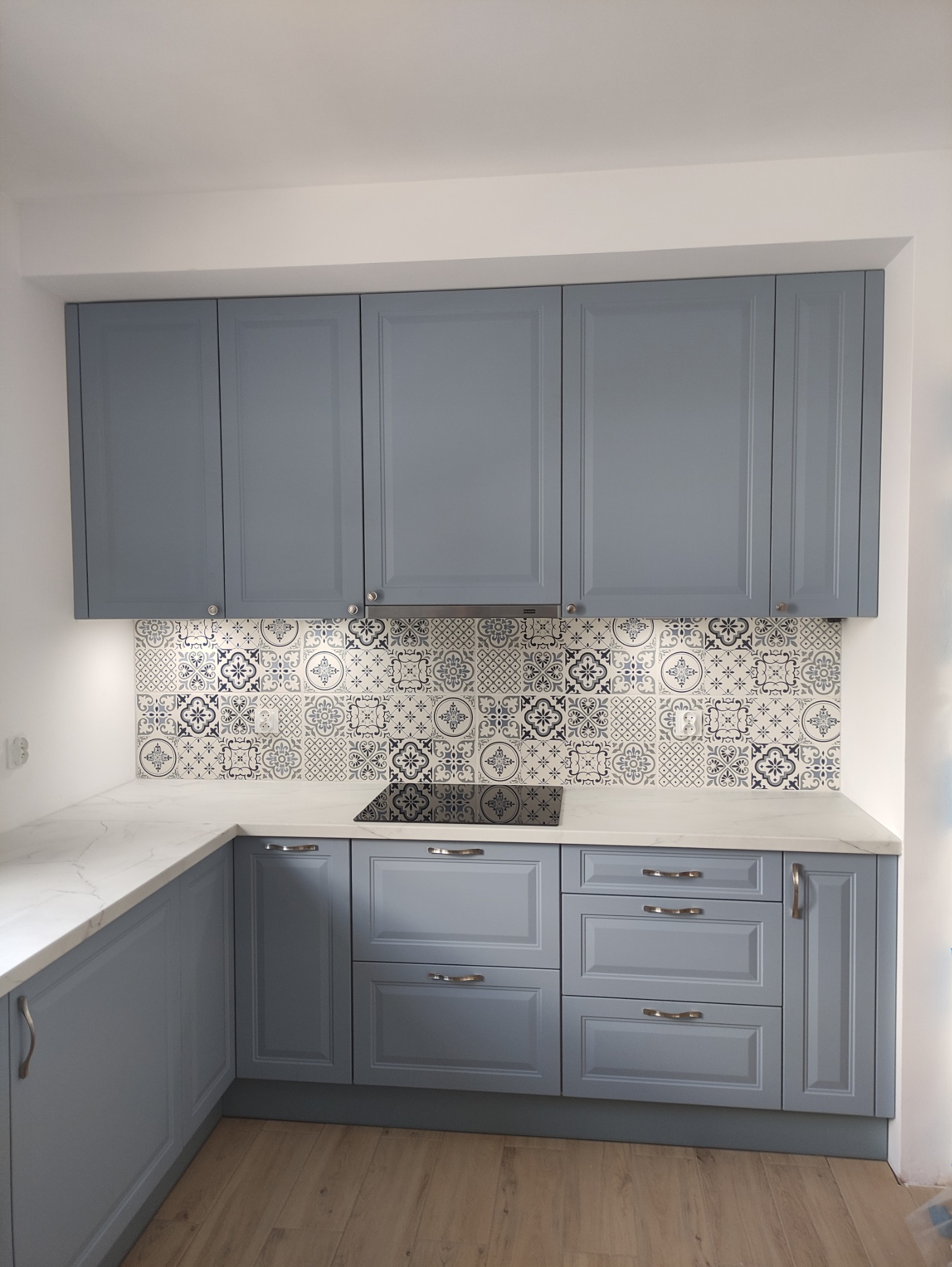 Niebieska zabudowa kuchenna z marmurowym blatem i dekoracyjnymi płytkami nad indukcją. Klasyczny styl, srebrne uchwyty, jasne drewno na podłodze.