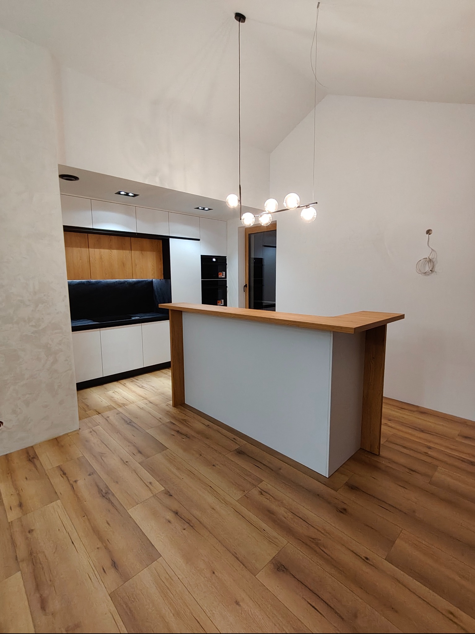 Nowoczesna kuchnia z białymi szafkami i drewnianym blatem, oświetlona wiszącą lampą. Podłoga z paneli imitujących drewno. Ściana z dekoracyjnym tynkiem.