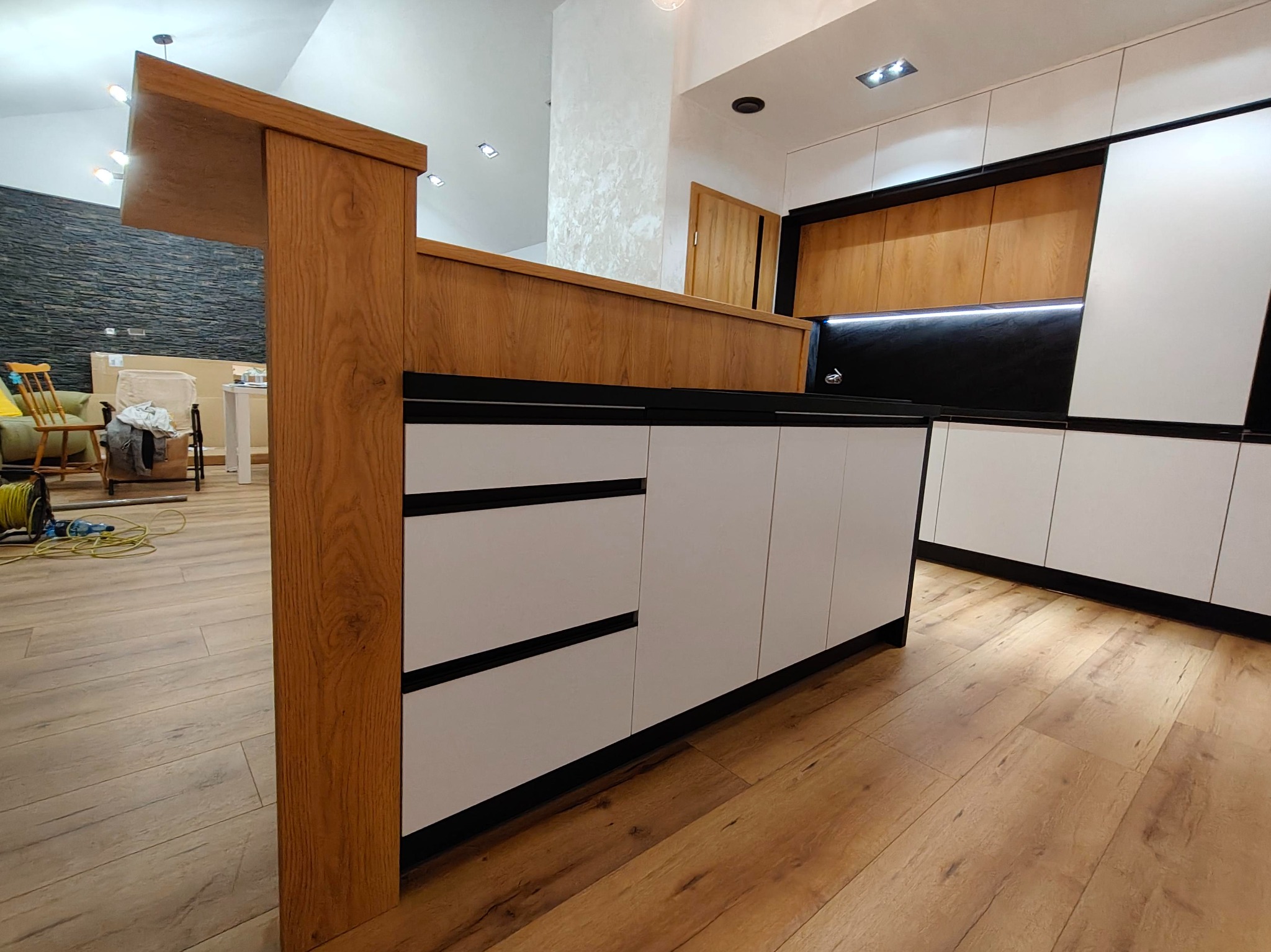 Nowoczesna kuchnia z białymi szafkami i drewnianym blatem, czarne akcenty, oświetlenie LED. Widoczna część wyspy kuchennej i drewniana podłoga.