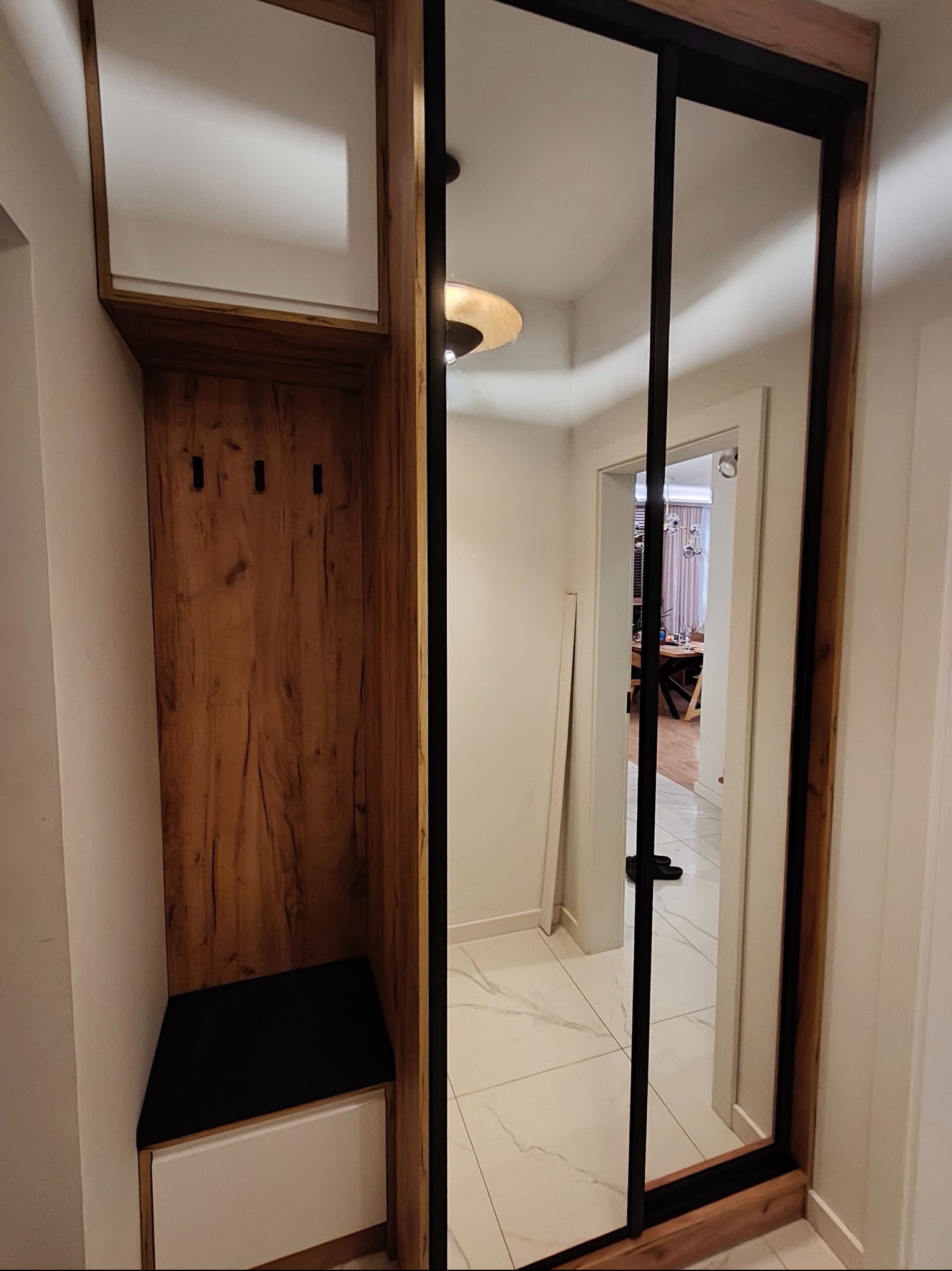 Funkcjonalna szafa wnękowa z lustrzanymi drzwiami przesuwnymi i drewnianym wykończeniem, z wbudowanym siedziskiem i wieszakami, optymalizująca przestrzeń w przedpokoju.