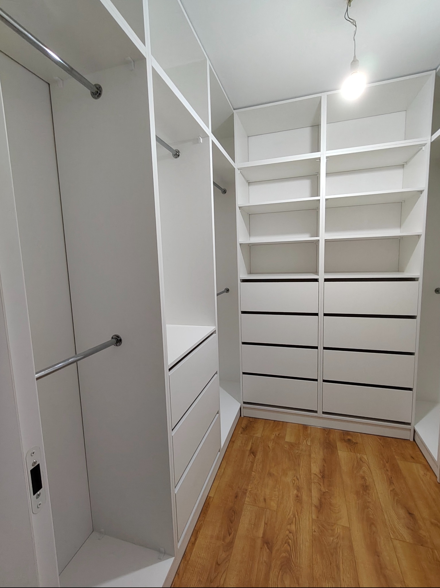 Biała, przestronna garderoba z półkami i szufladami, widok z perspektywy wnętrza. Chromowane drążki na ubrania i ciepła podłoga z paneli dodają elegancji.