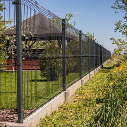 EK-BUD II sp z o.o. - Długi, panelowy płot w kolorze grafitowym, z betonową podmurówką, oddzielający posesję z altaną od łąki z żółtymi kwiatami w słoneczny dzień.