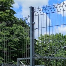 EK-BUD II sp z o.o. - Nowoczesne, szare ogrodzenie panelowe z metalu na tle zieleni drzew i błękitnego nieba. Widoczne detale mocowania paneli. Minimalistyczny design.