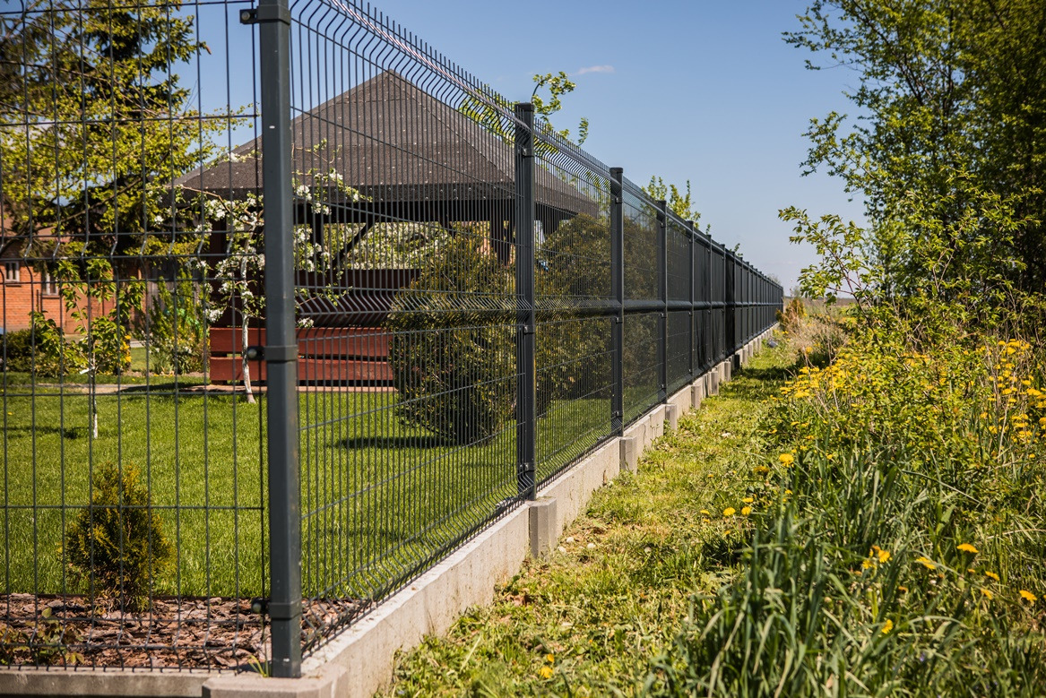 Długi, panelowy płot w kolorze grafitowym, z betonową podmurówką, oddzielający posesję z altaną od łąki z żółtymi kwiatami w słoneczny dzień.