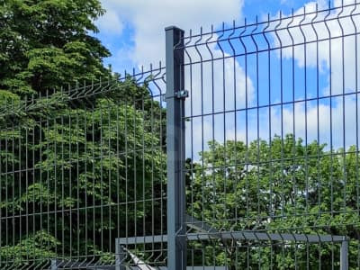 Nowoczesne, szare ogrodzenie panelowe z metalu na tle zieleni drzew i błękitnego nieba. Widoczne detale mocowania paneli. Minimalistyczny design.