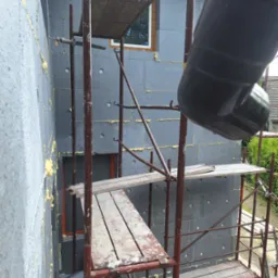 Ocieplony szary styropian, rusztowanie z drewnianymi podestami, fragment rynny. Prace na wysokości, widok z dołu na elewację budynku.