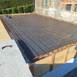 X-Men building academy - Zbrojenie stropu na drewnianym szalunku, widok z góry. Siatka z prętów stalowych ułożona na czarnej folii. W tle mur z cegły i zielony żywopłot.