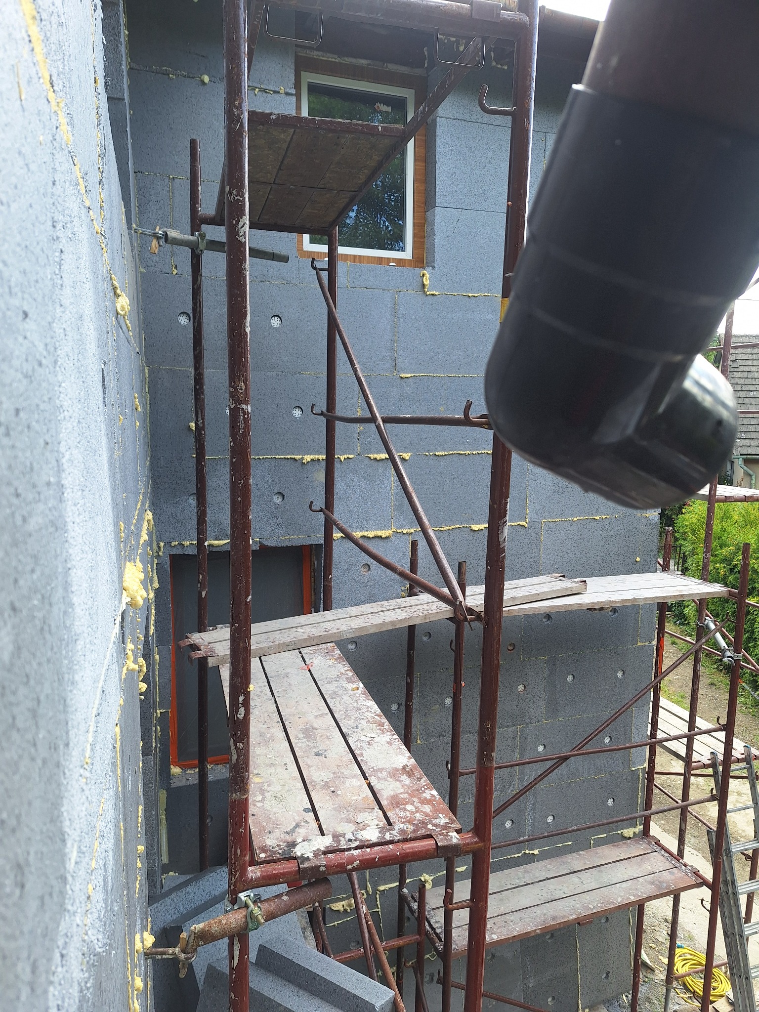 Ocieplony szary styropian, rusztowanie z drewnianymi podestami, fragment rynny. Prace na wysokości, widok z dołu na elewację budynku.