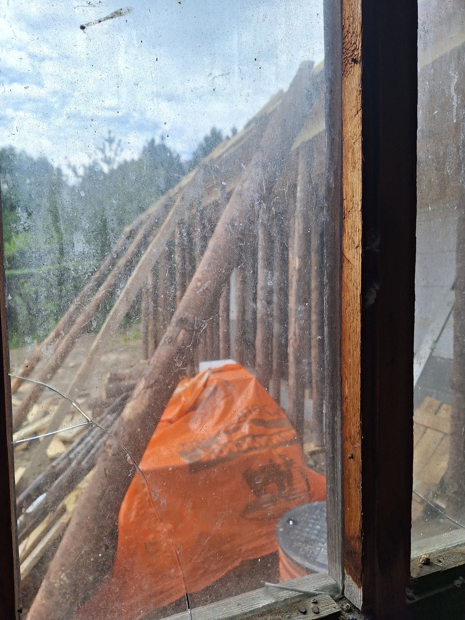 Konstrukcja szkieletowa domu w budowie, widoczna przez brudne, pęknięte okno. Na pierwszym planie drewniana rama okienna i pomarańczowa plandeka.