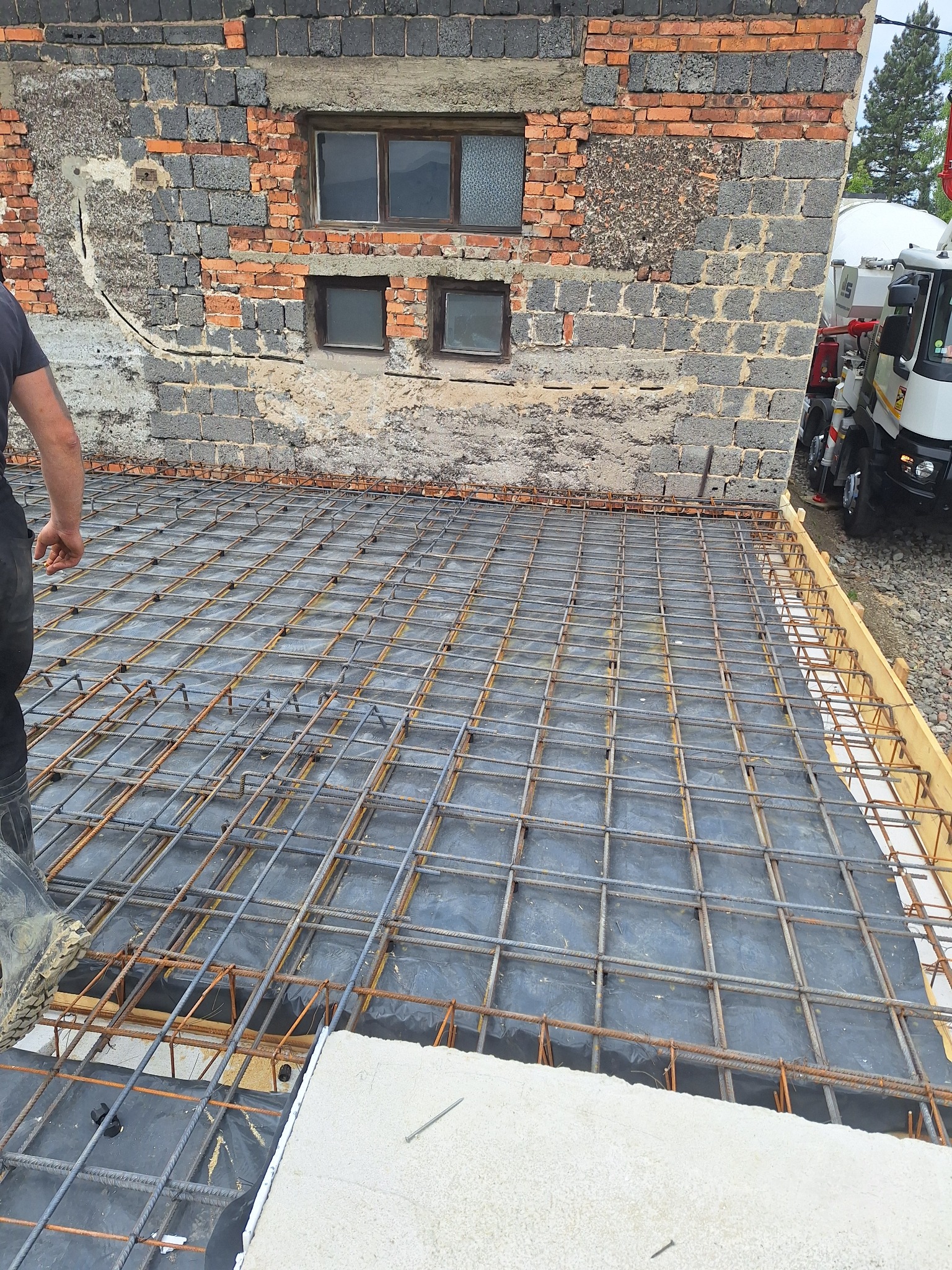 Zbrojenie pod wylewkę fundamentu na czarnej folii, widoczna ściana budynku z cegły oraz betoniarka w tle. Przygotowanie do betonowania.