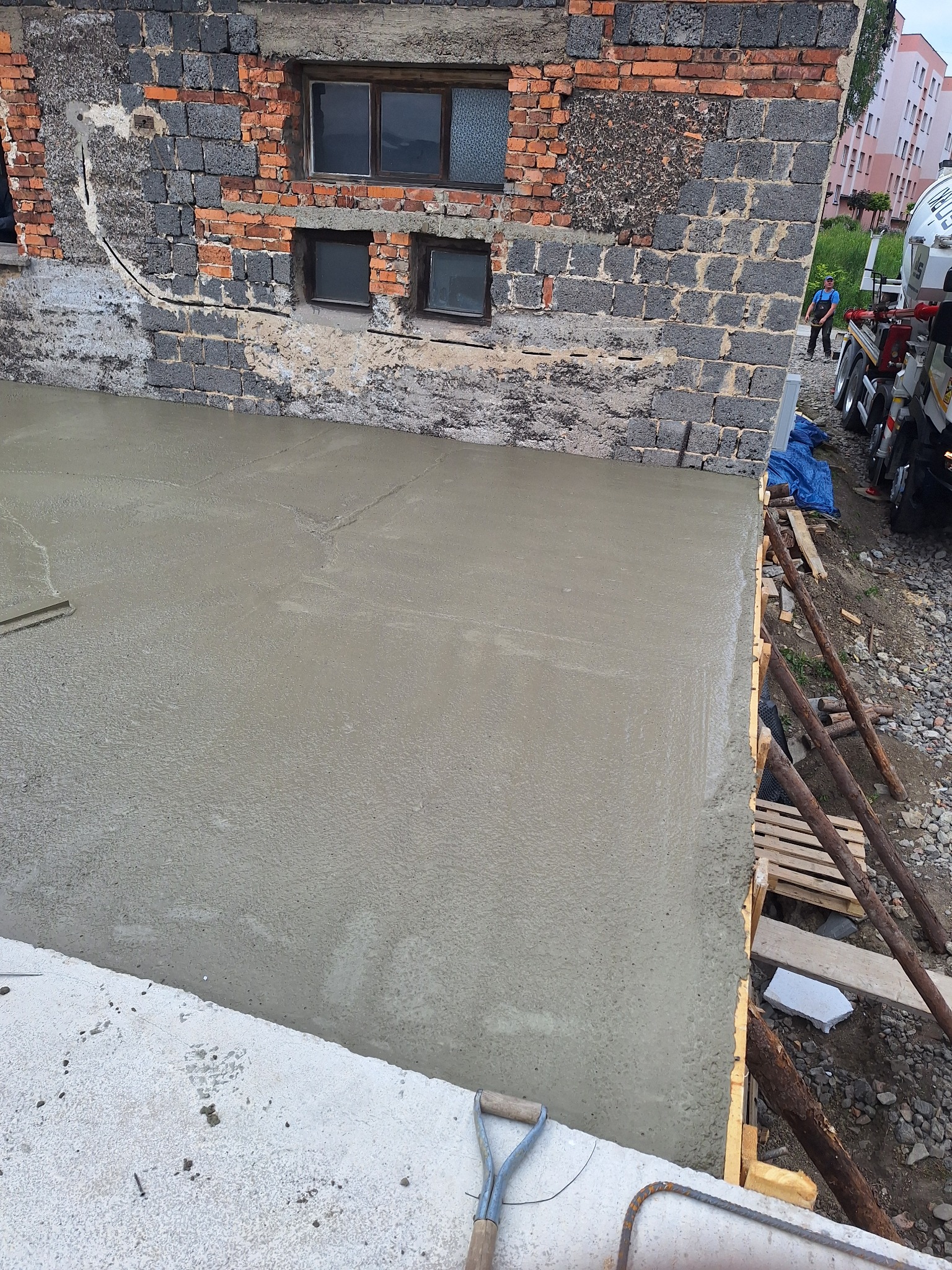 Wylany beton przy starej, ceglanej ścianie budynku z oknami. Widoczne szalunki, narzędzia i betoniarka w tle. Prace budowlane w toku.