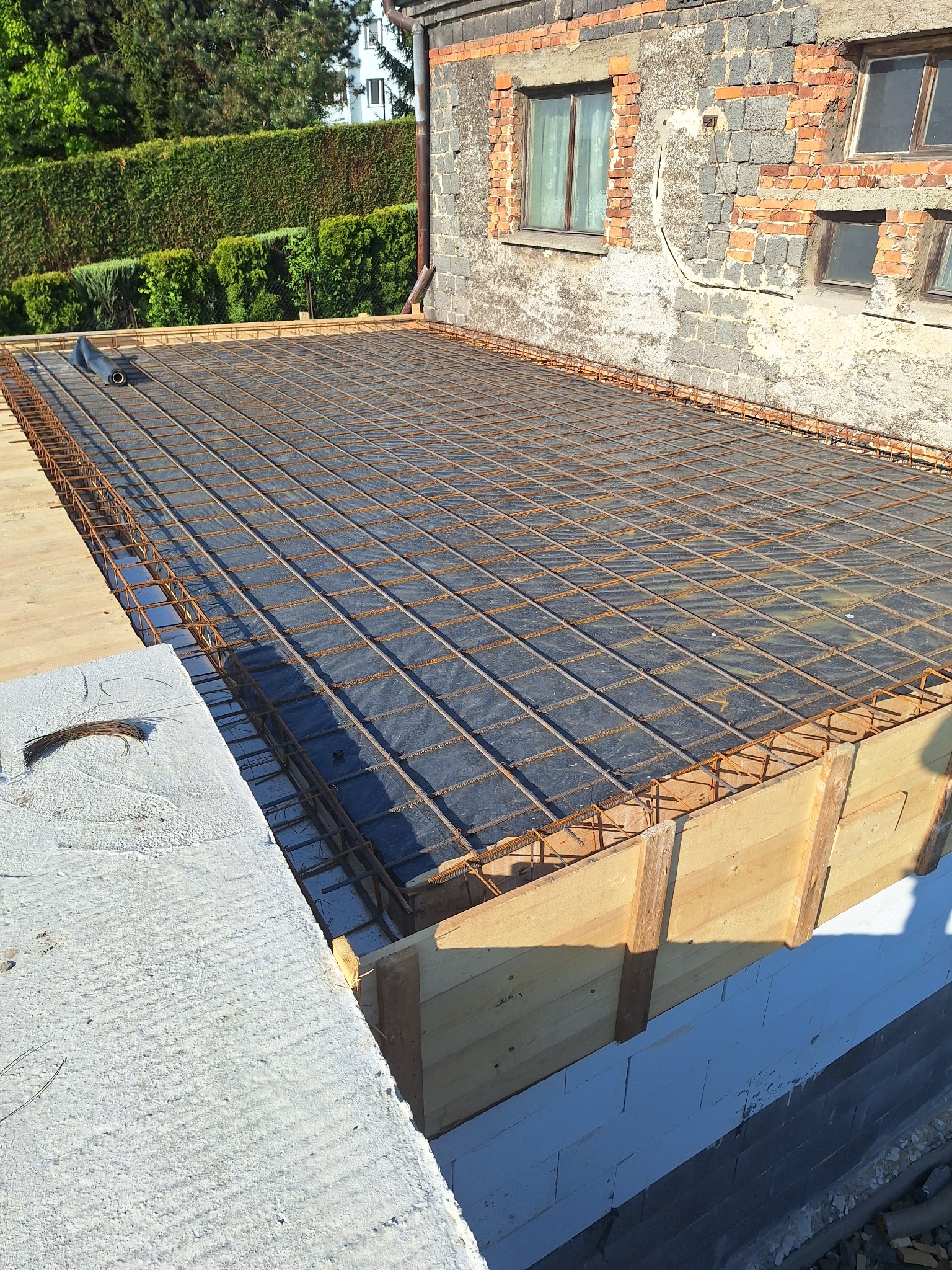 Zbrojenie stropu na drewnianym szalunku, widok z góry. Siatka z prętów stalowych ułożona na czarnej folii. W tle mur z cegły i zielony żywopłot.