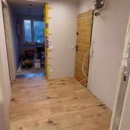 MAR-BUS SPÓŁKA Z OGRANICZONĄ ODPOWIEDZIALNOŚCIĄ - Wnętrze w trakcie remontu: Ułożona podłoga z paneli imitujących drewno, widoczna ościeżnica, niezabezpieczona instalacja elektryczna i poziomica na ścianie przy wejściu do kolejnego pomieszczenia.