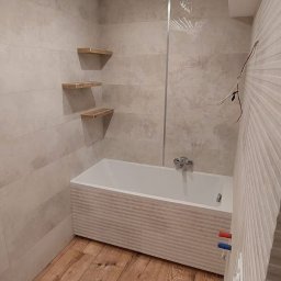MAR-BUS SPÓŁKA Z OGRANICZONĄ ODPOWIEDZIALNOŚCIĄ - Nowoczesna łazienka z drewnianą podłogą, beżowymi płytkami imitującymi kamień, białą wanną z poziomymi żłobieniami i trzema drewnianymi półkami na ścianie.