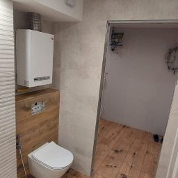 MAR-BUS SPÓŁKA Z OGRANICZONĄ ODPOWIEDZIALNOŚCIĄ - Nowoczesna łazienka z wiszącą toaletą, grzejnikiem i drewnopodobną podłogą, widok na sąsiednie pomieszczenie z niedokończonymi pracami elektrycznymi.